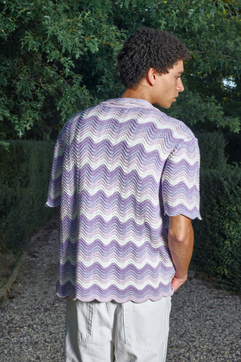 Person wearing a purple and white patterned shirt standing outdoors with greenery in the background