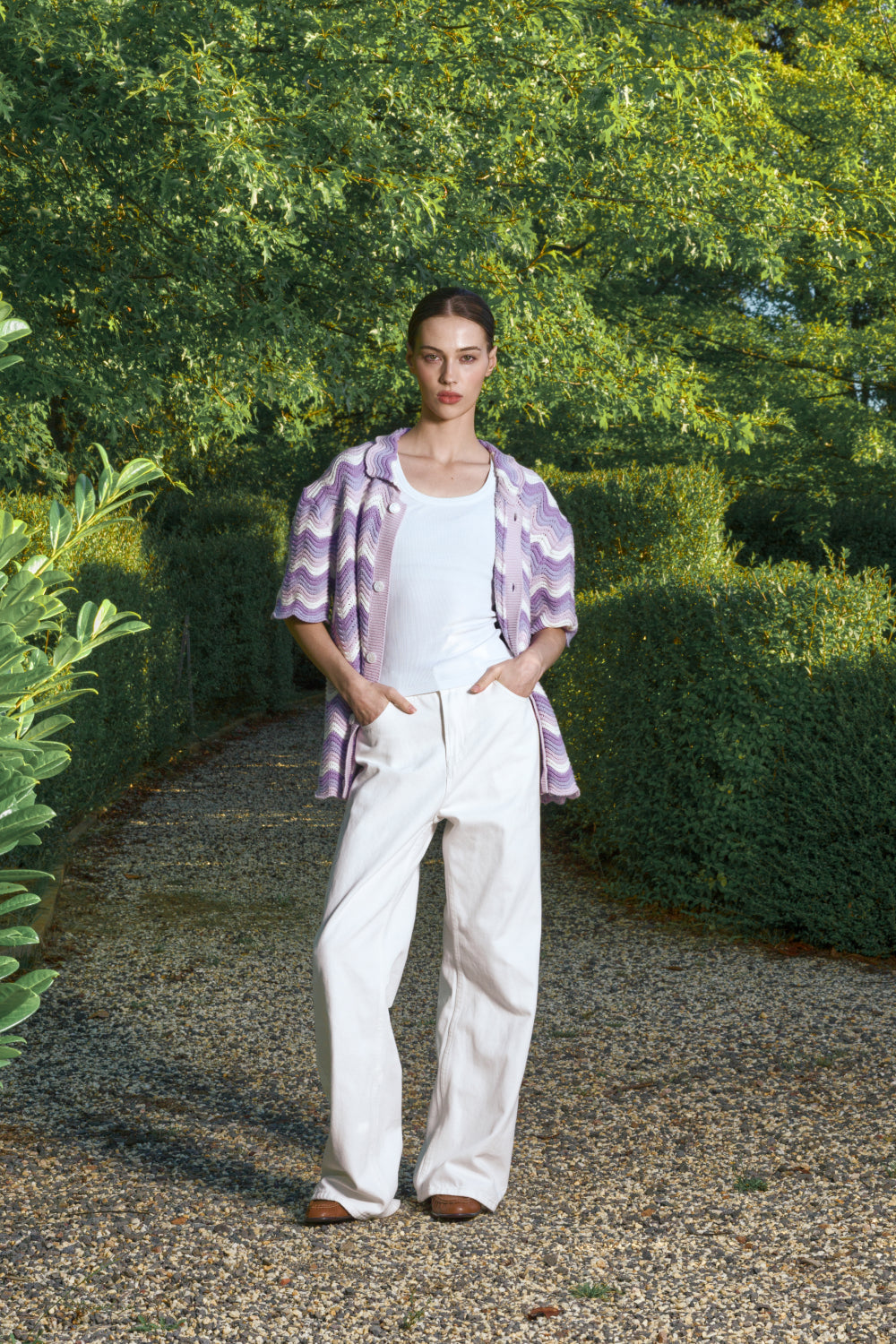 Woman standing in a garden wearing a purple patterned shirt and white pants.