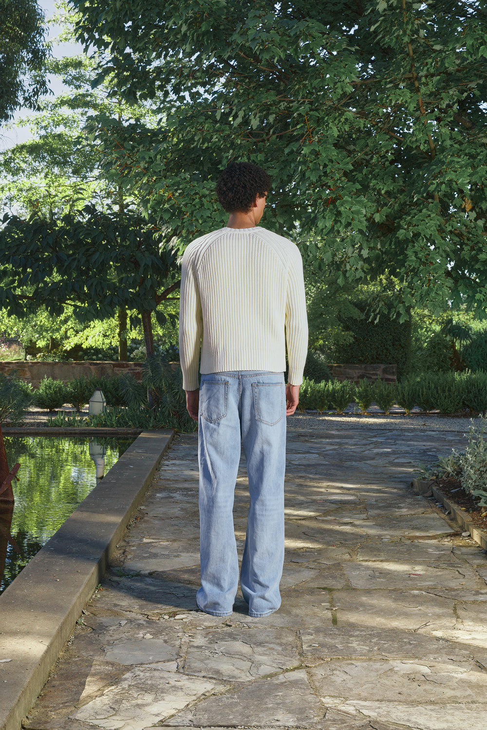 Back view for a model wearing a cream ribbed knit sweater with light jeans, standing outdoors.