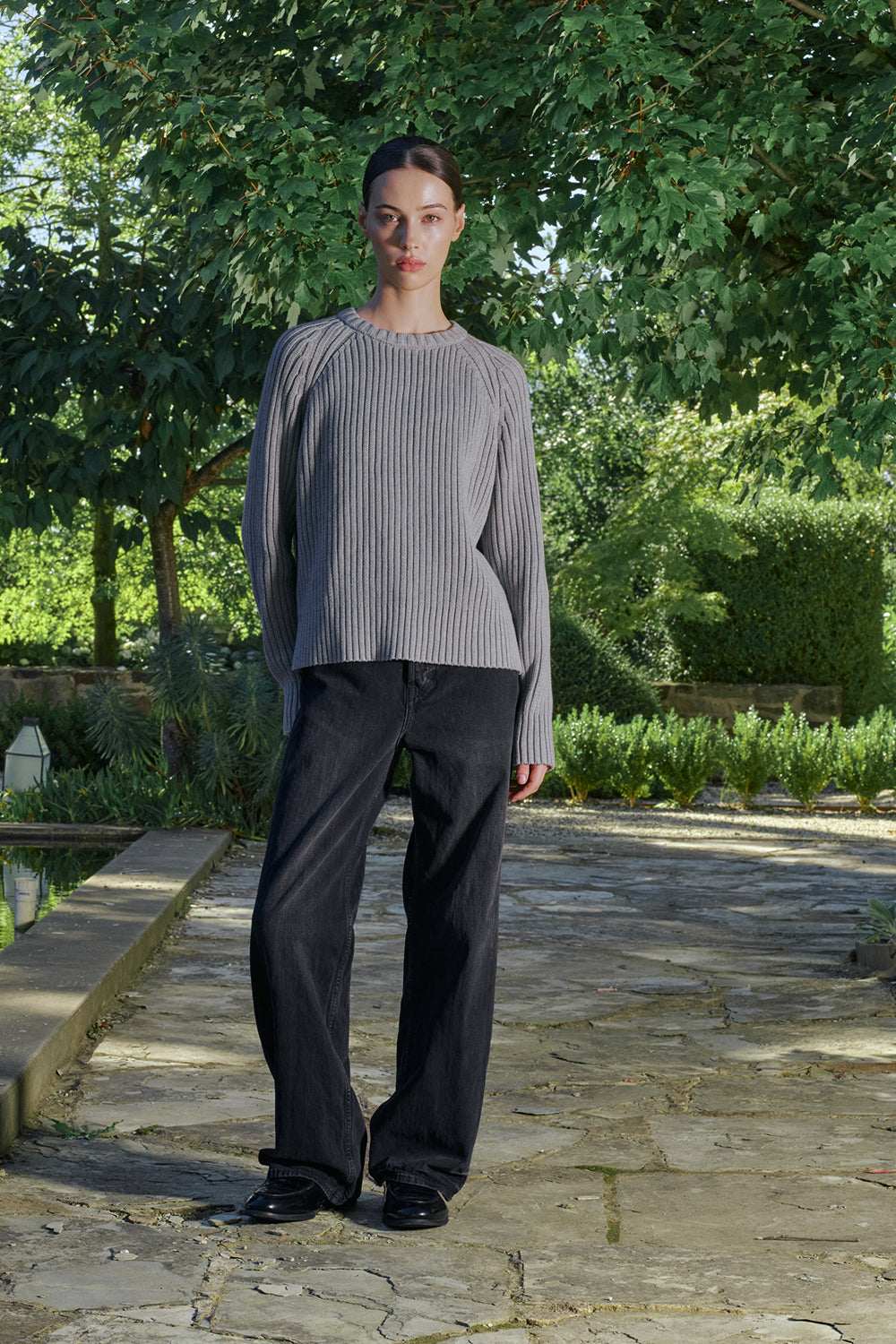 Model wearing a grey ribbed knit sweater with black pants, standing outdoors.
