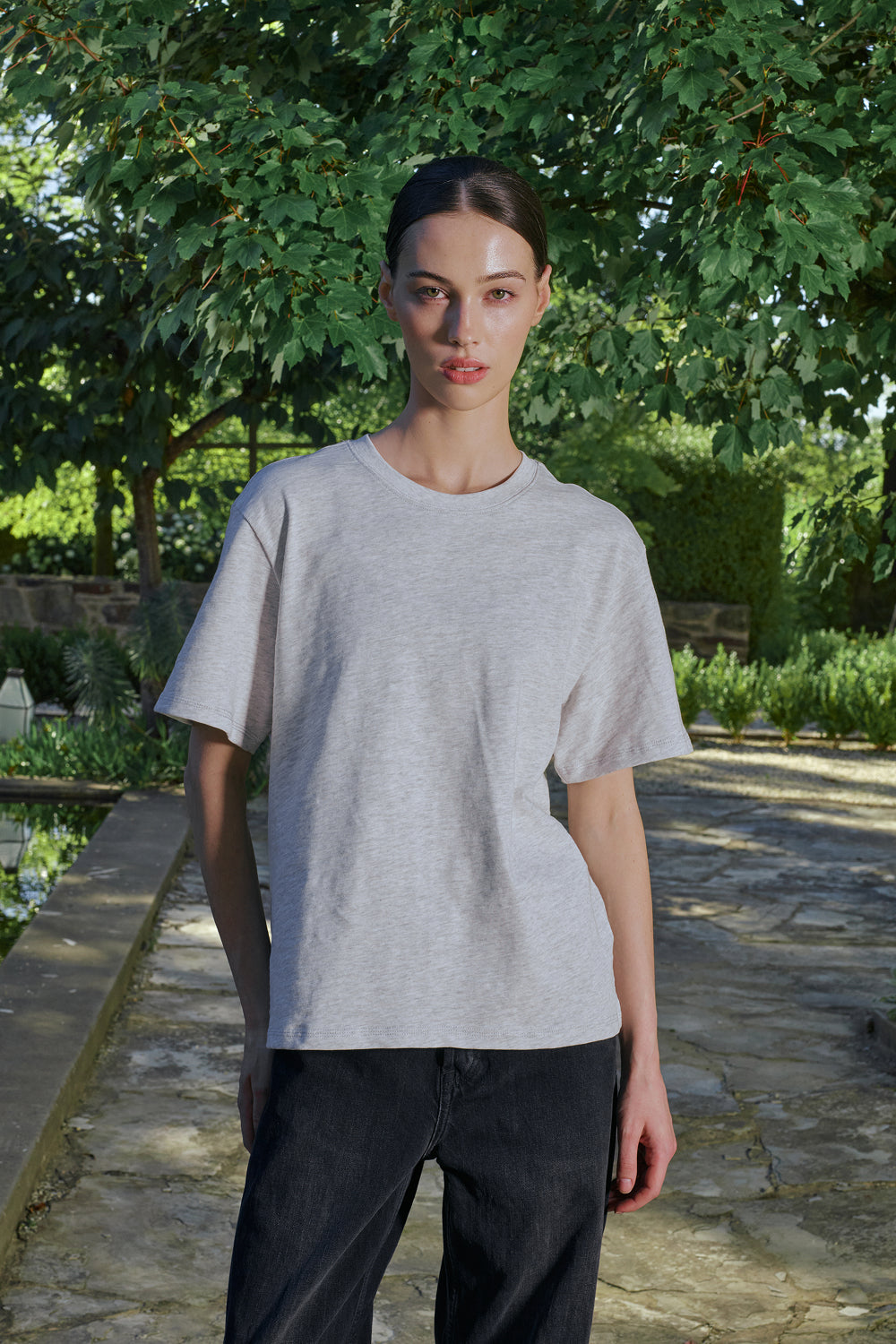 Person wearing a gray t-shirt and dark pants standing outdoors with greenery in the background