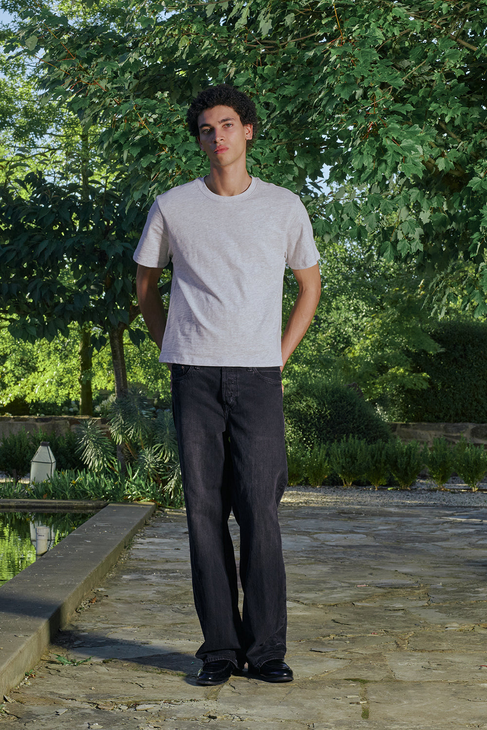 Model wearing a gray t-shirt and dark pants standing outdoors.