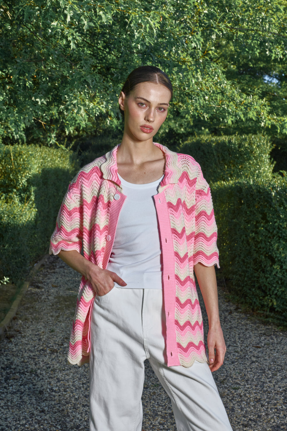 Woman wearing a pink and cream shale stitch crochet shirt over a white top and white pants, standing outdoors with greenery in the background.