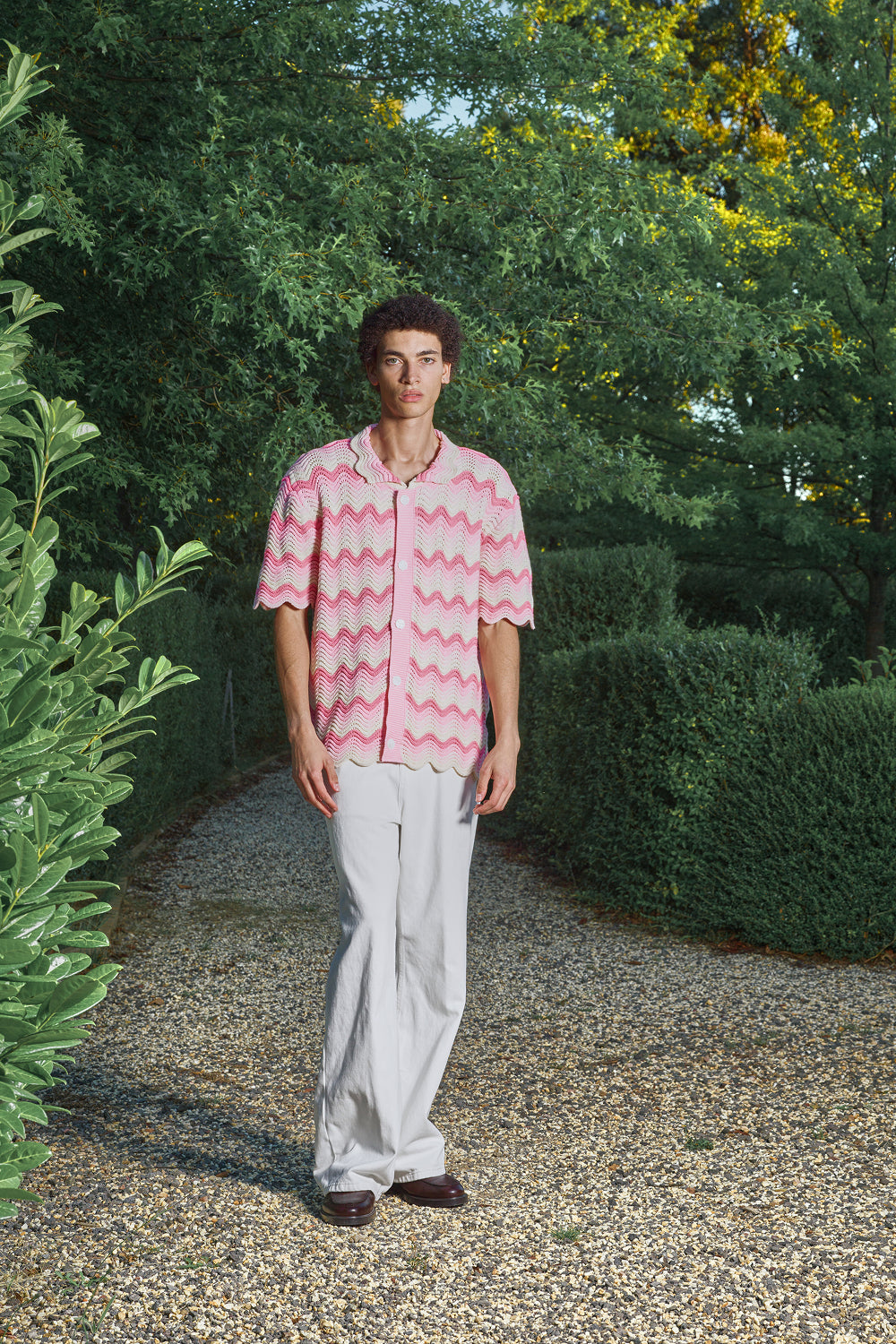 Man wearing a pink shale stitch pattern shirt and white pants standing in a garden.