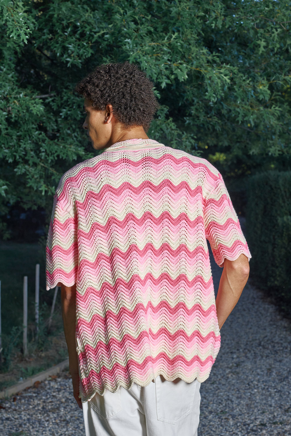 Model wearing a pink and cream shale stitch shirt standing outdoors with greenery in the background
