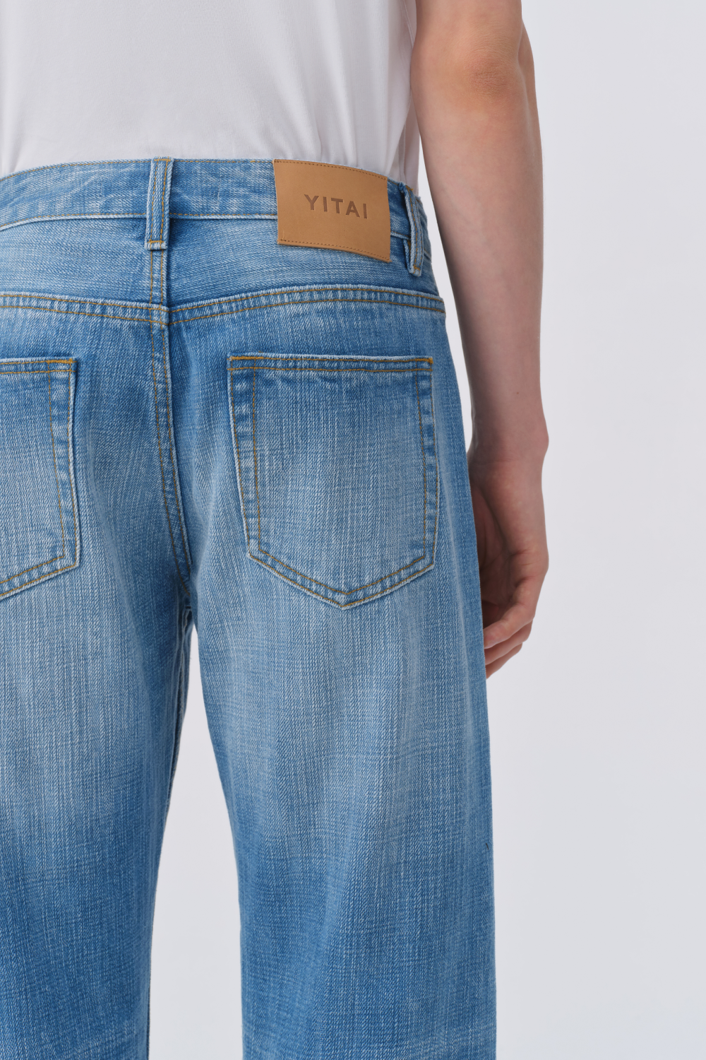 Close-up of the back of light blue denim trousers, showing the back patch pockets and YITAI leather waistband patch.