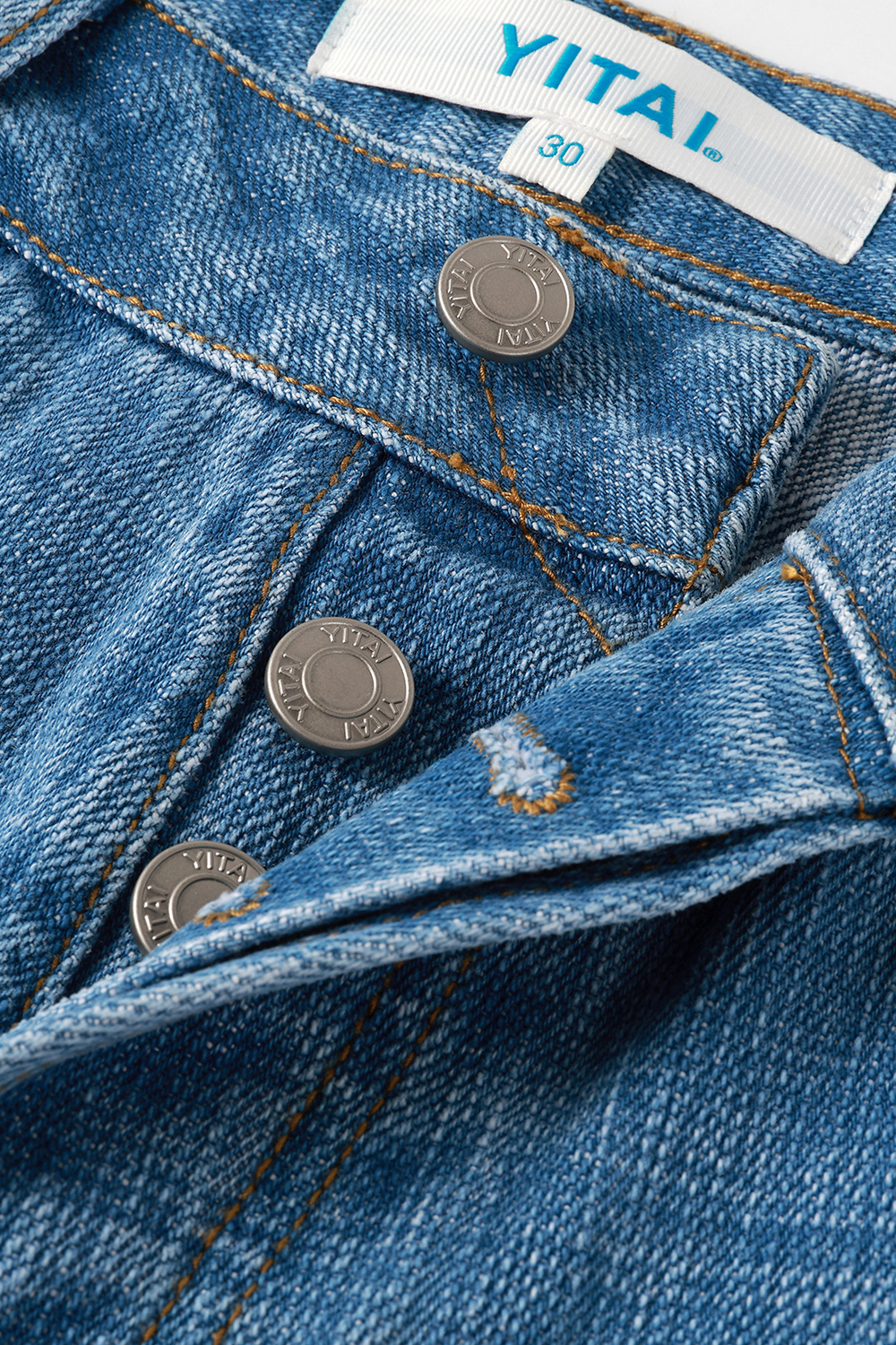 Close-up of light blue denim trousers showing the button fly with engraved YITAI metal buttons and the interior size label.