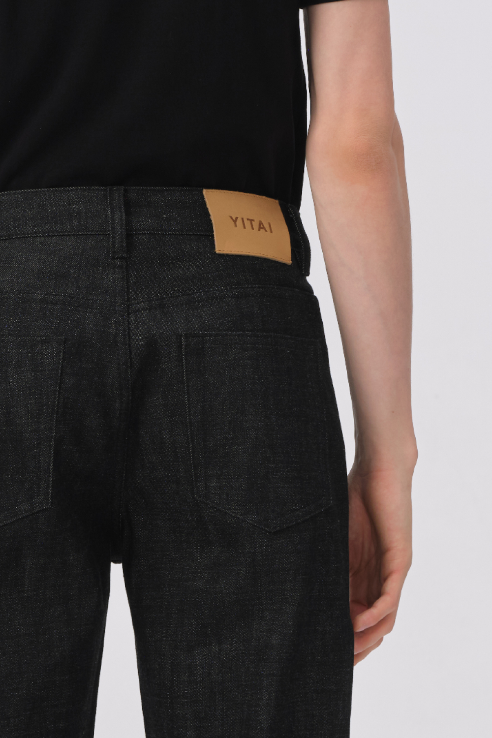 Close-up of the back of black denim trousers, showing the back patch pockets and YITAI leather waistband patch.