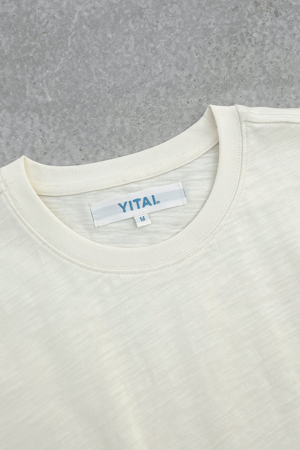 Close-up of a cream crewneck T-shirt laid flat on a gray textured surface, featuring a white neck label with blue “YITAI.” branding and size M tag.