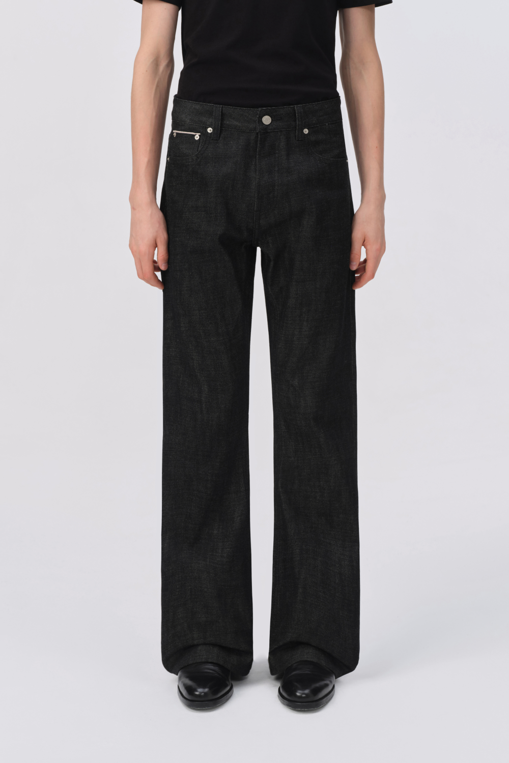 Close-up of black raw denim jeans on a model, showing the button fly, front pockets, and straight-leg silhouette.