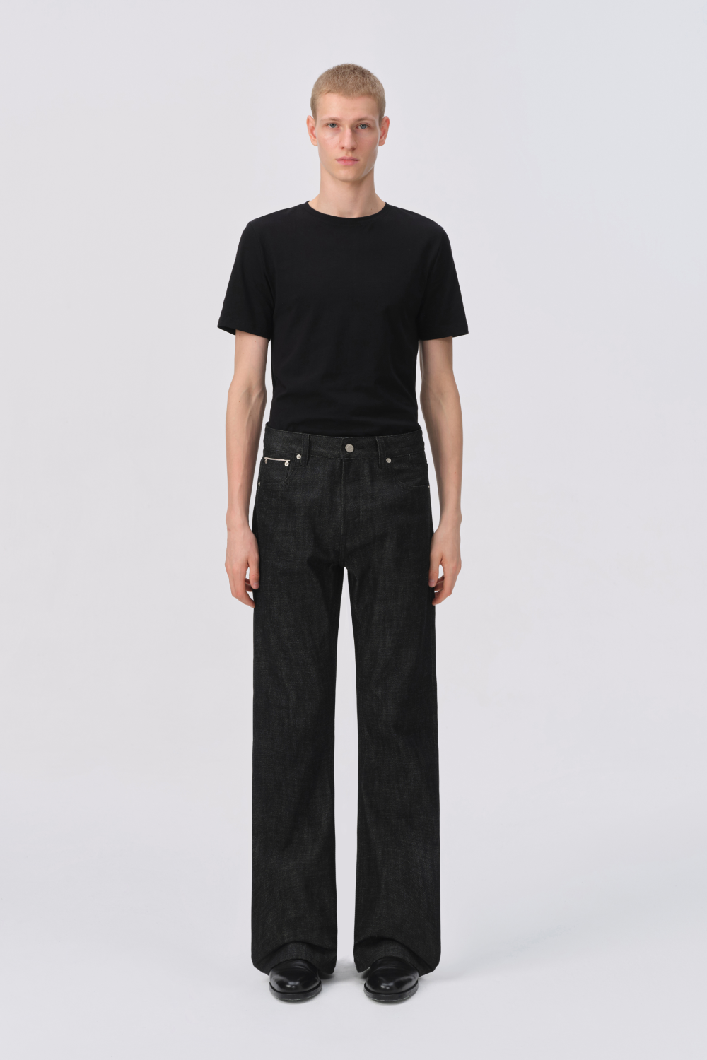 Model wearing a black t-shirt with black raw denim, straight-leg jeans in a relaxed fit, styled with black shoes.