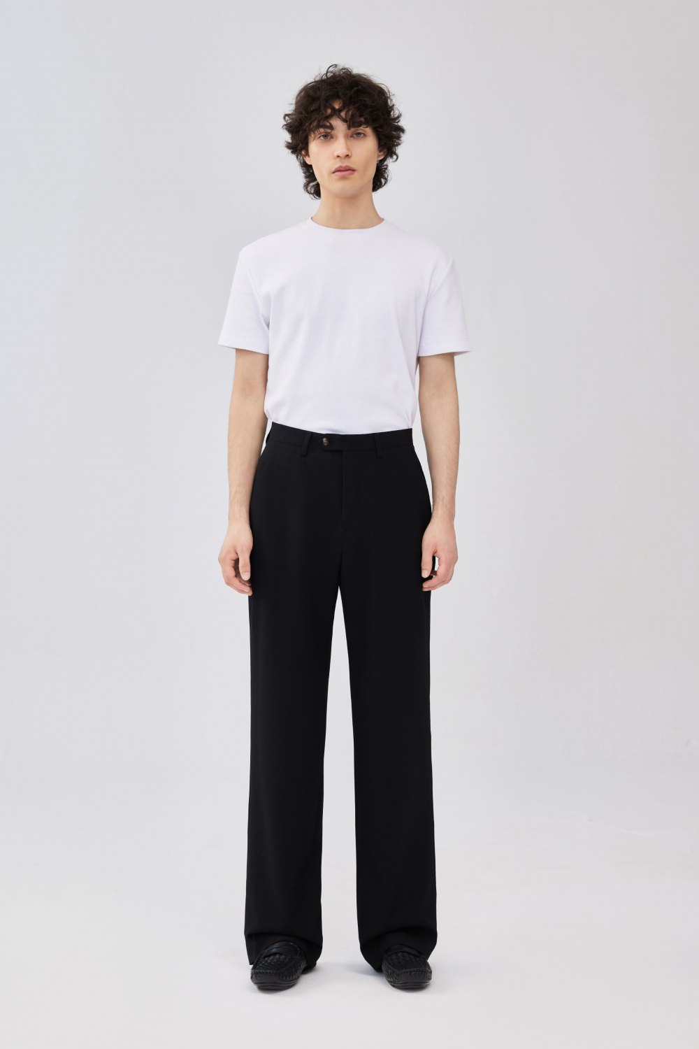 Full-length front view of a model wearing black wool trousers with a straight leg, front crease, belt loops, side pockets, and button closure, styled with a white T-shirt.
