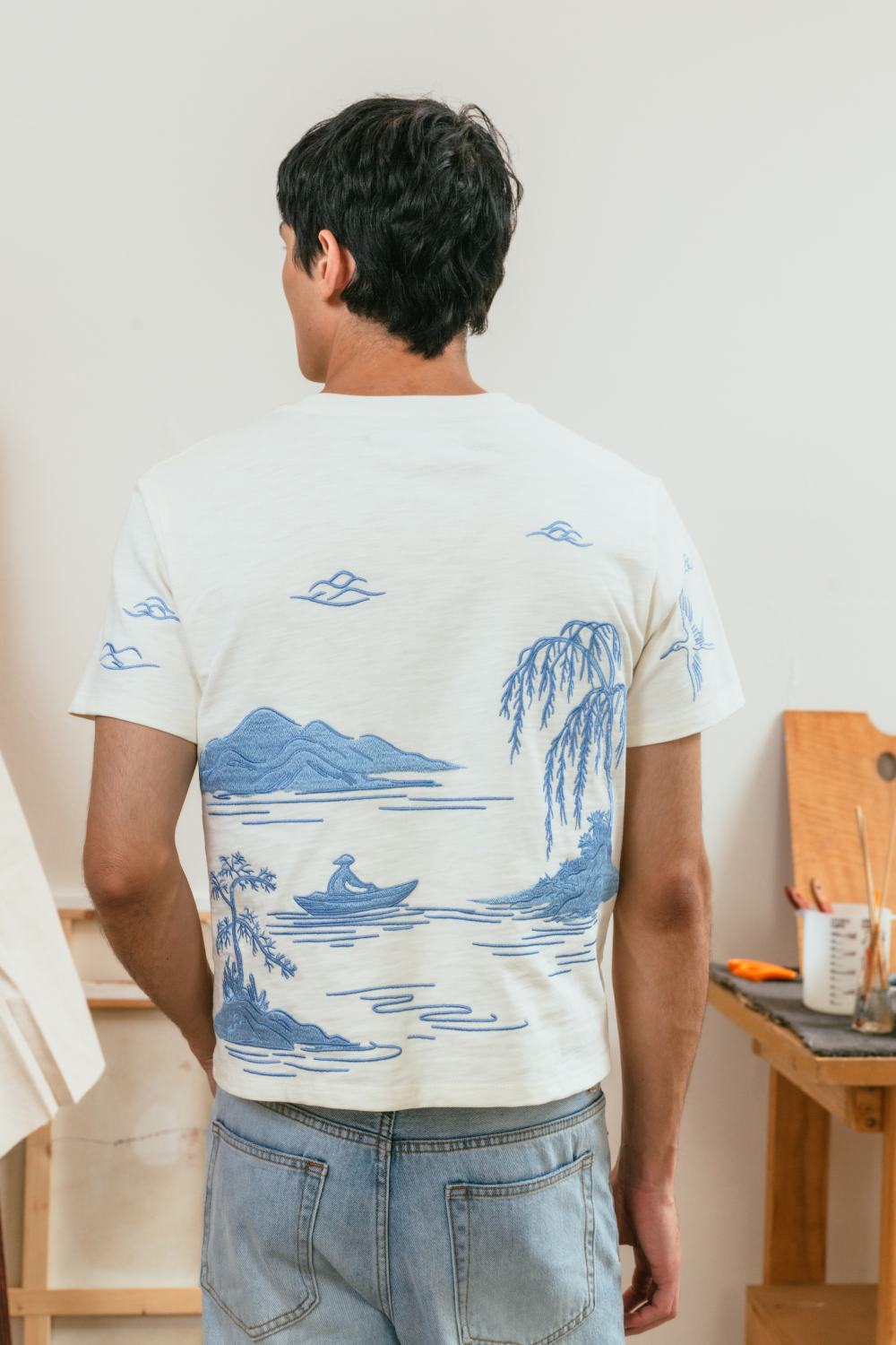Back view of a person wearing a white t-shirt with blue nature-themed embroidery, standing in a room.