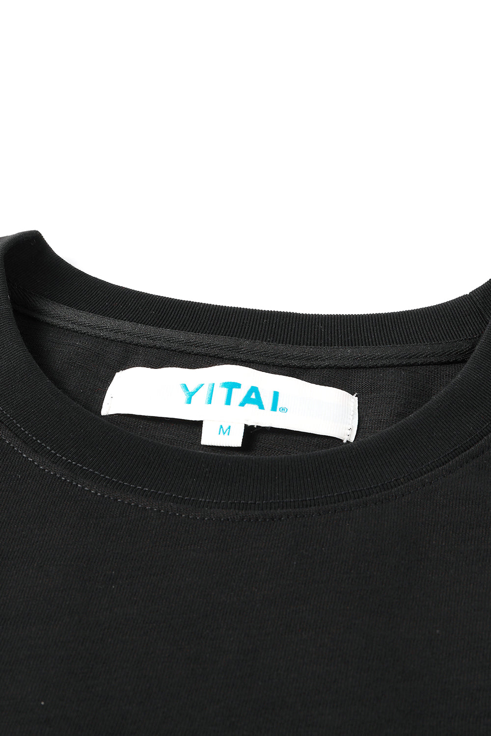 Close-up of a Black crewneck T-shirt laid flat on a gray textured surface, featuring a white neck label with blue “YITAI.” branding and size M tag.