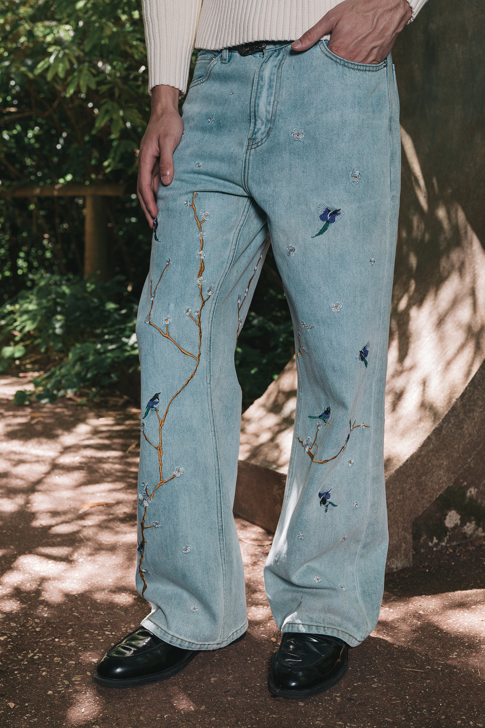 Light blue jeans with tree branch and bird embroidery worn by a person outdoors.