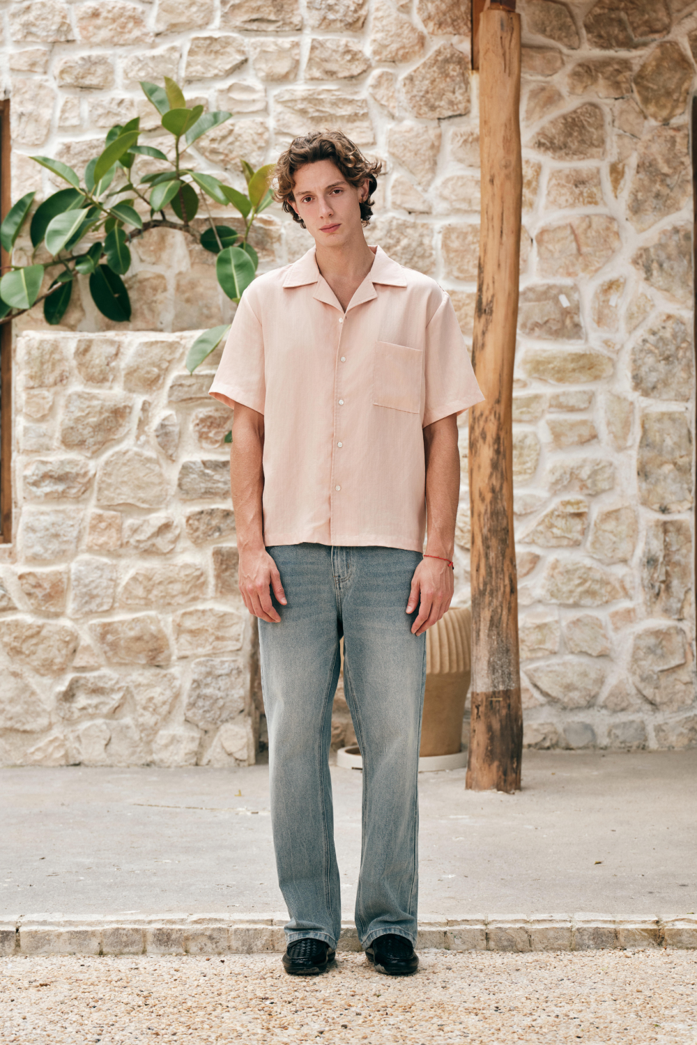 Full length view of a model wearing a light pink short-sleeve button-up shirt with a camp collar and chest patch pocket, styled with a light blue jeans.