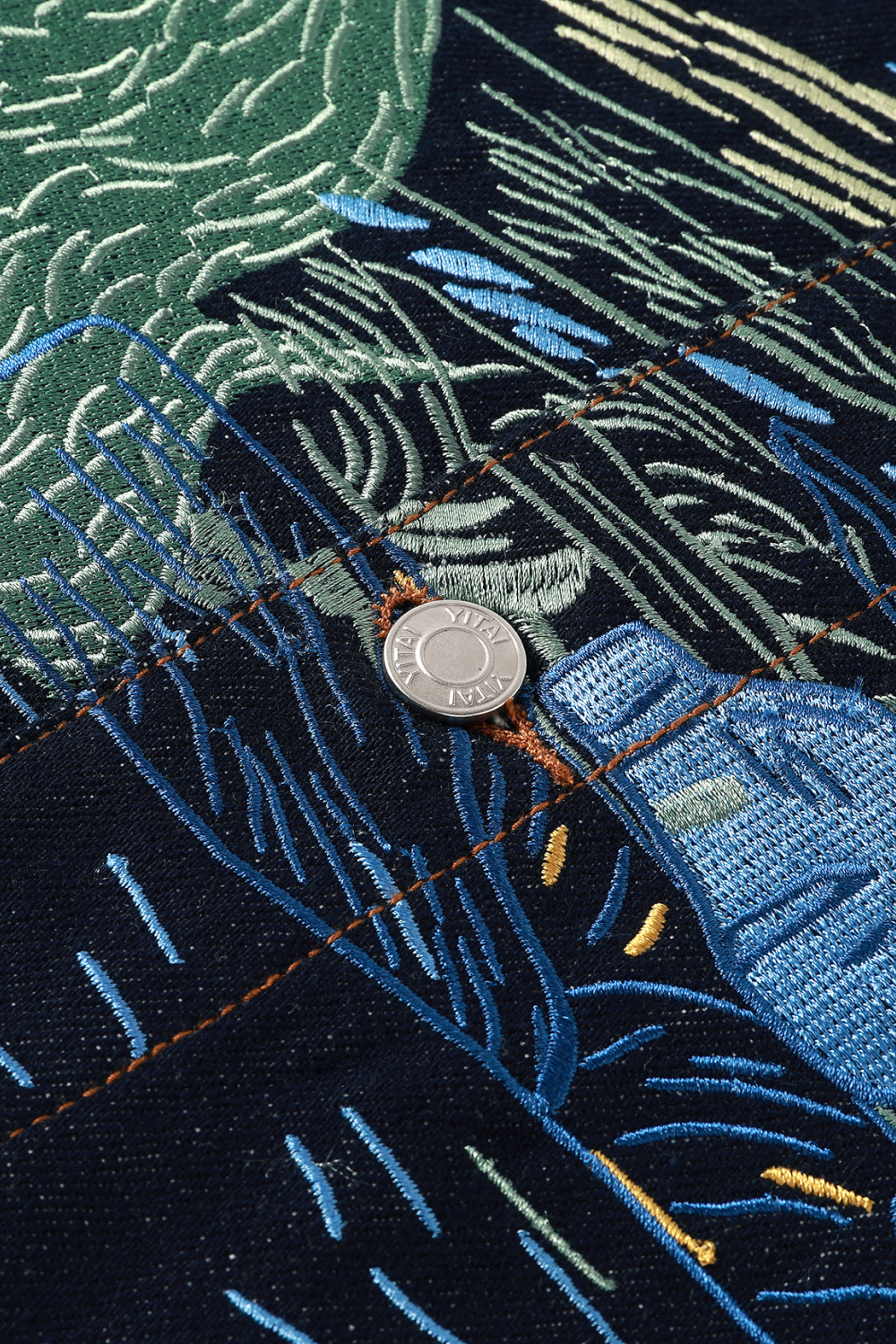 Close-up of dark denim with Starry Night-inspired embroidery in shades of blue, green, yellow, and cream, featuring detailed stitching and a silver button.
