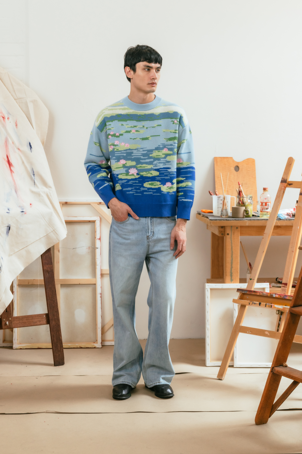 Full-body image of a model wearing the blue lily pond sweater with light wash jeans in an artist studio setting.