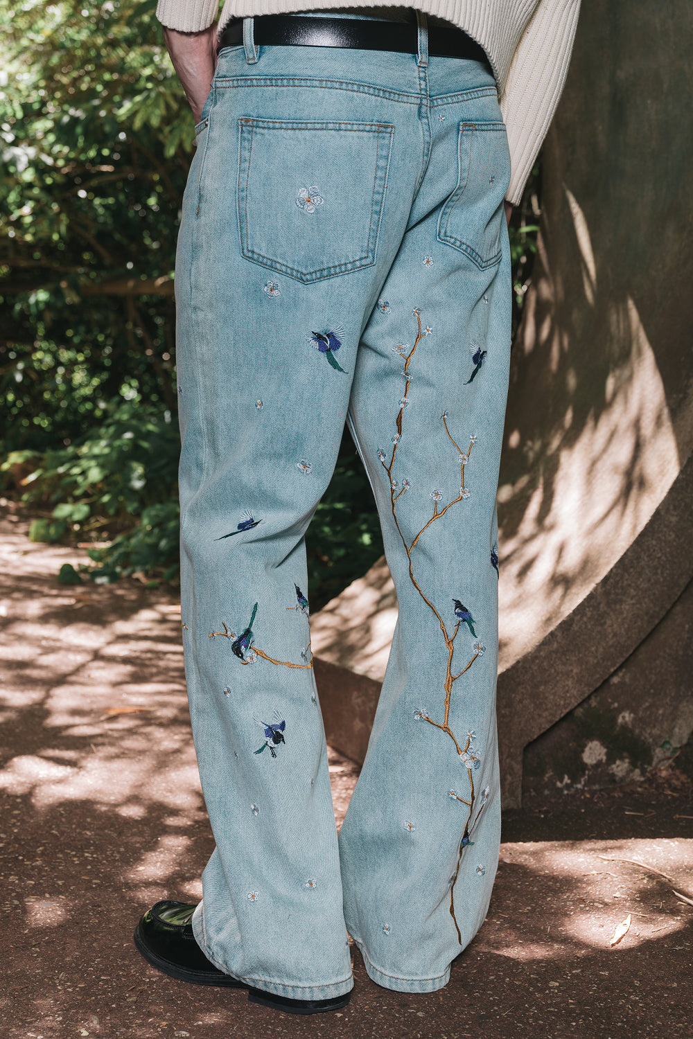 Back view of model wearing light blue denim jeans with bird and branch embroidery
