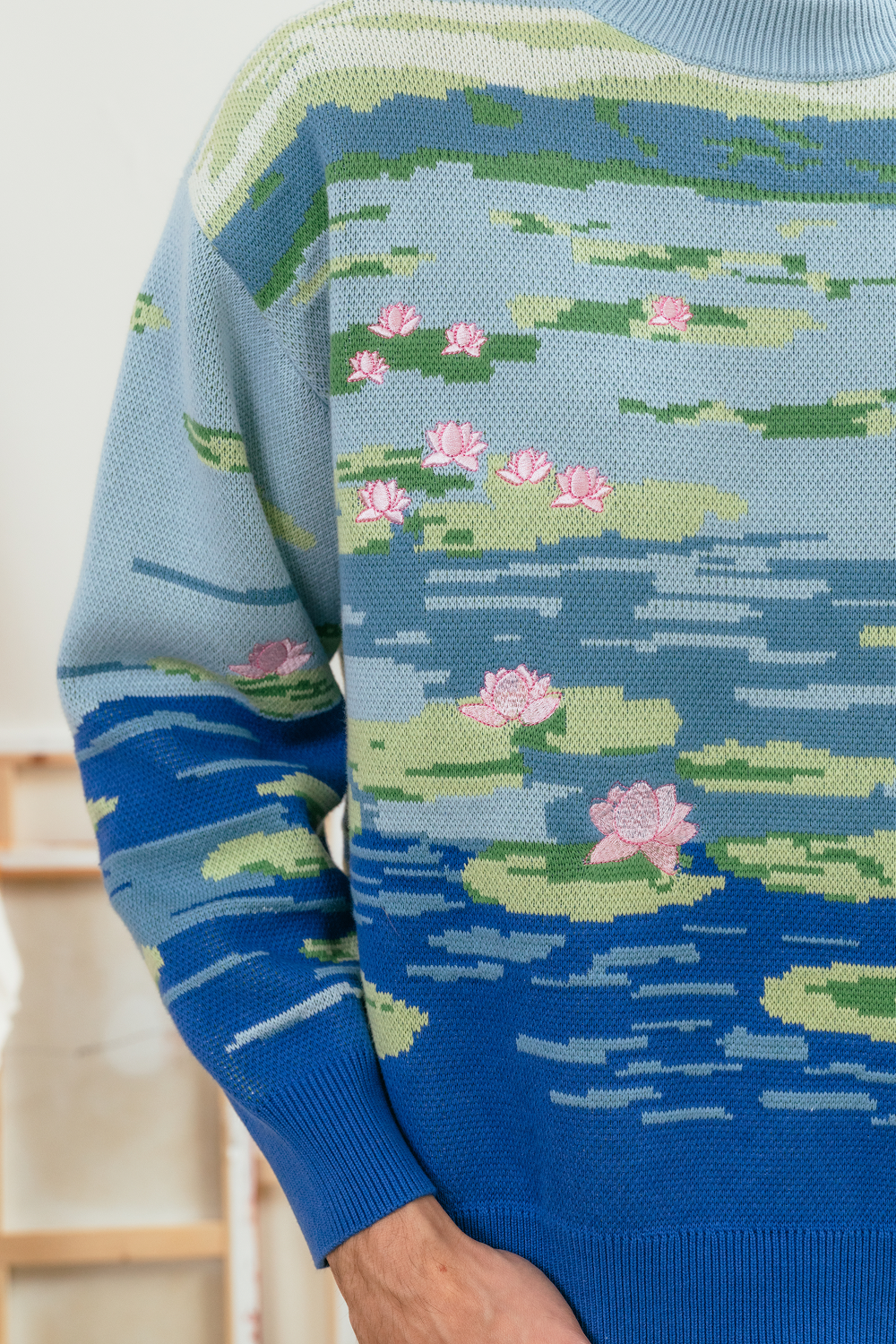 Close-up of a light blue knit sweater featuring a lily pond jacquard design with green lily pads, layered blue water tones, and pink lotus flower embroidery across the front and sleeve.
