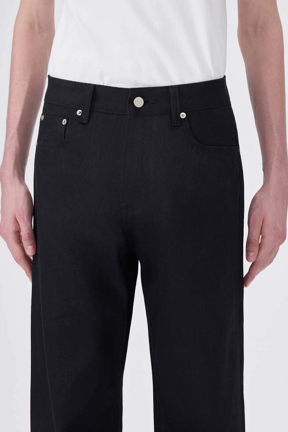 Close-up front view of black selvedge denim jeans showing the waistband, silver button, rivets, and front pockets.