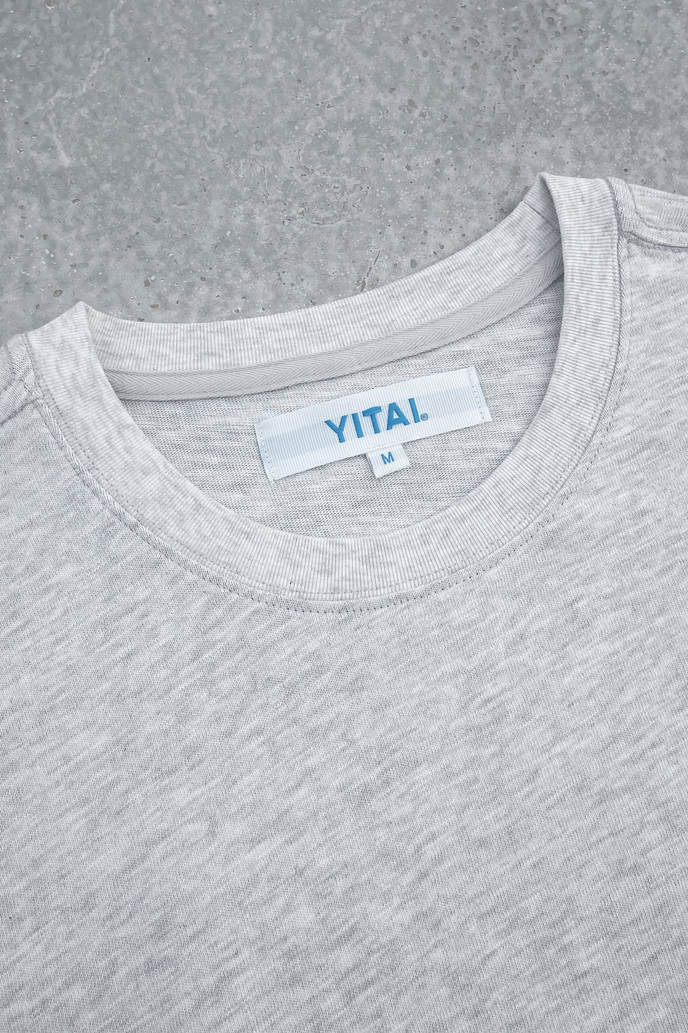 Close-up of a light heather gray crewneck T-shirt laid flat on a gray textured surface, featuring a white neck label with blue “YITAI.” branding and size M tag.
