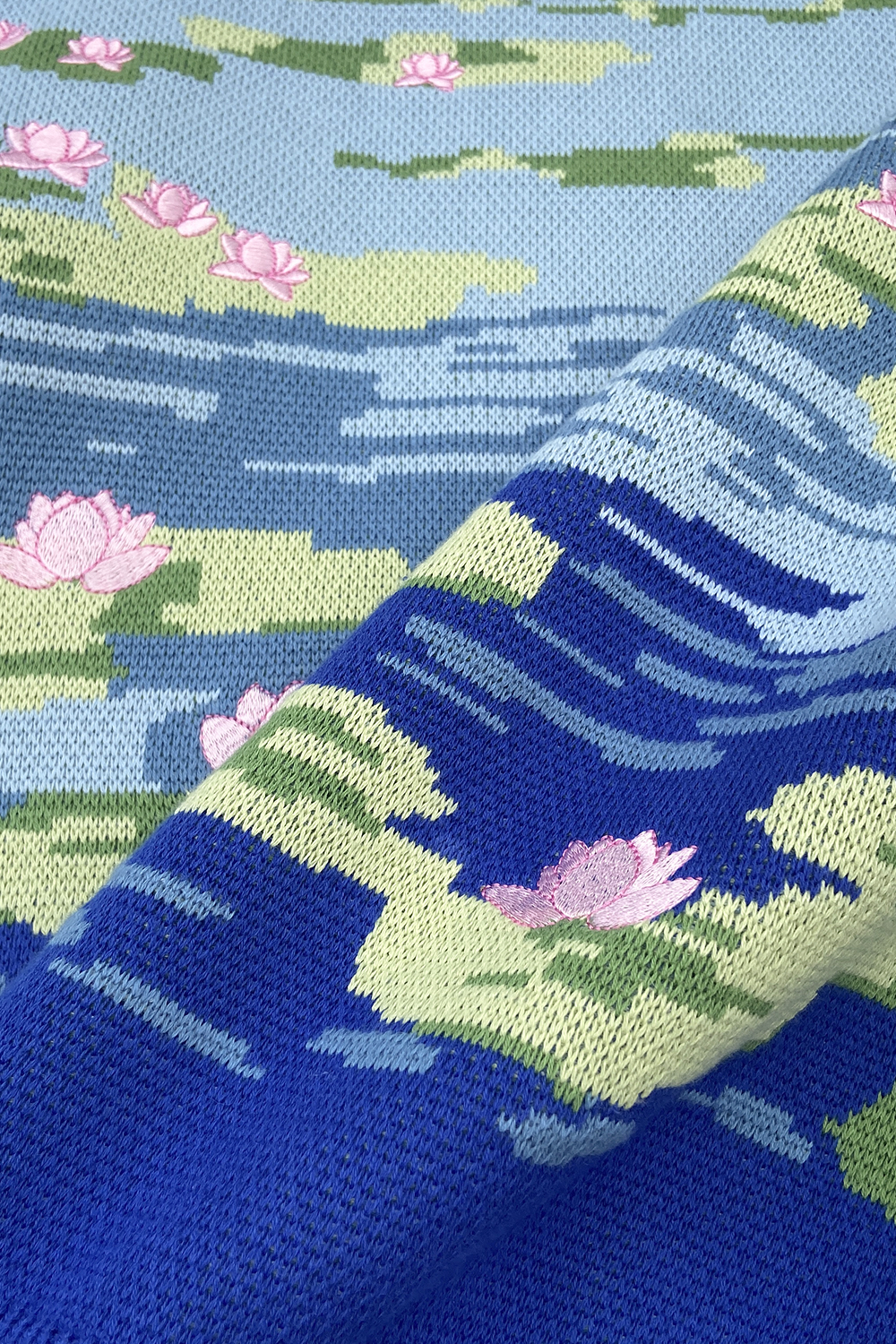 Product-only image of the light blue sweater featuring a lily pond jacquard knit design with floating lily pads, pink lotus flowers, and deep blue water detailing near the hem.