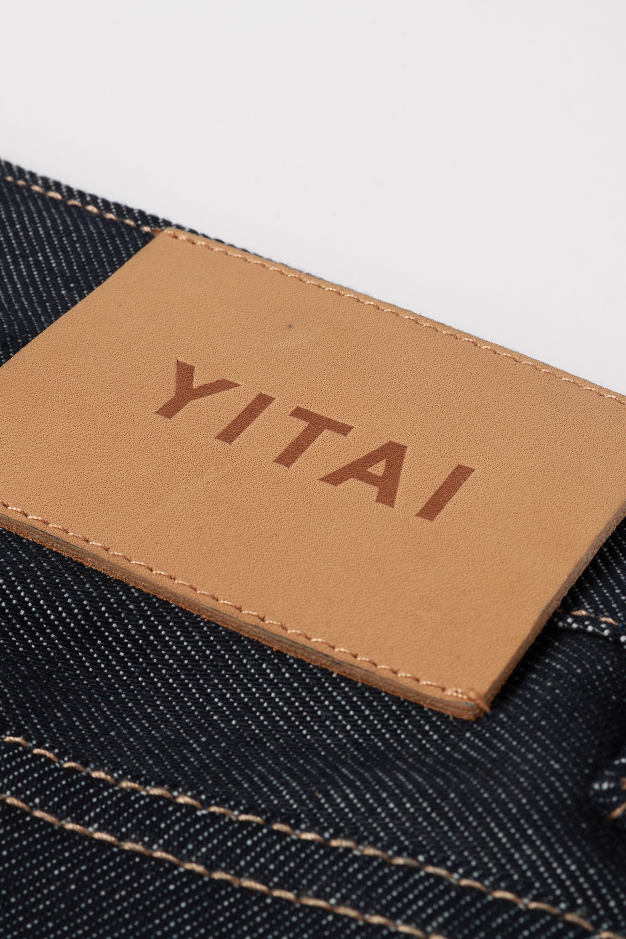 Close-up of a tan leather brand patch with the word 'YITAI' printed in brown, attached to dark blue denim with contrast stitching.
