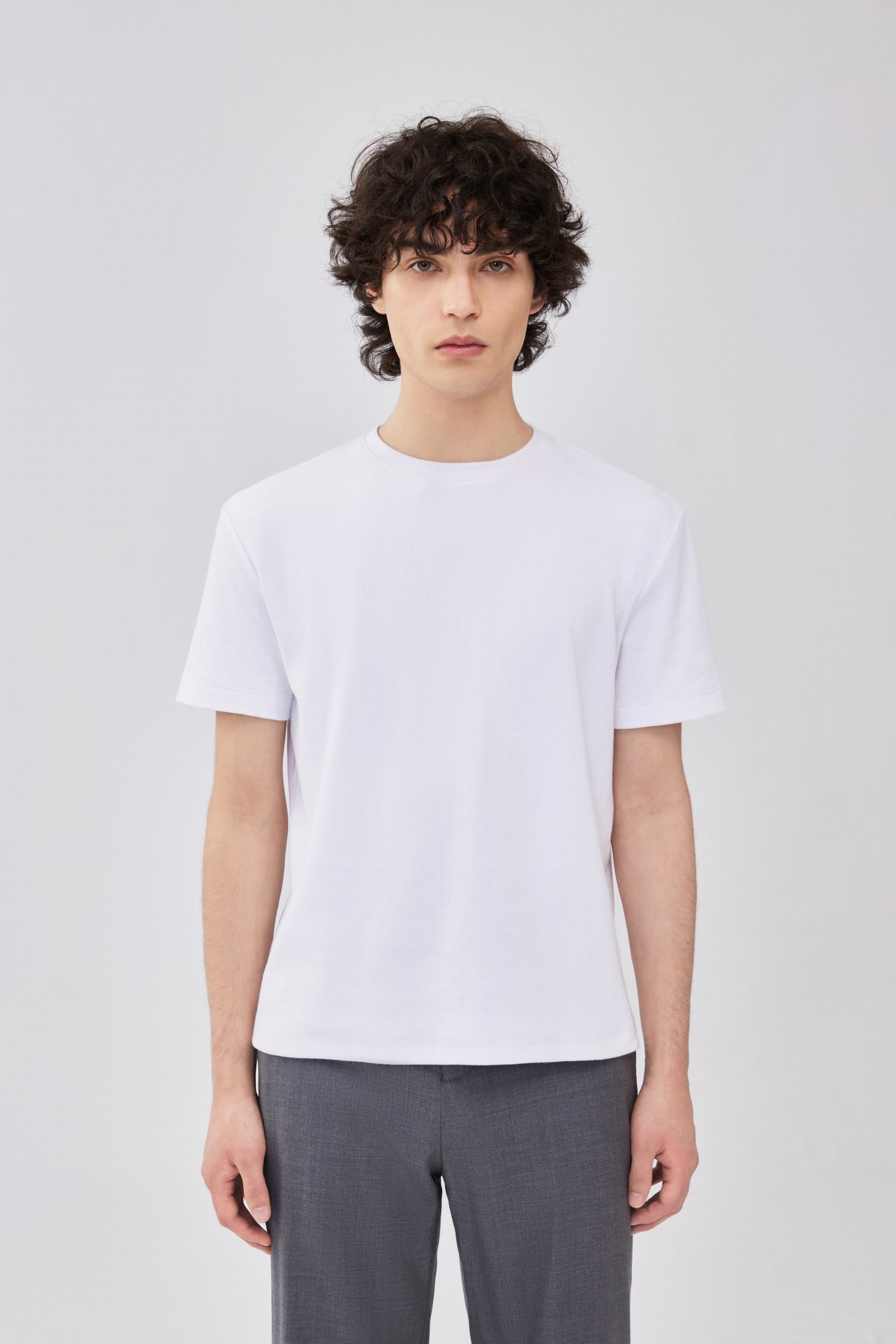 A male model wearing a plain white short-sleeve crewneck t-shirt and gray trousers, standing against a light gray studio background.