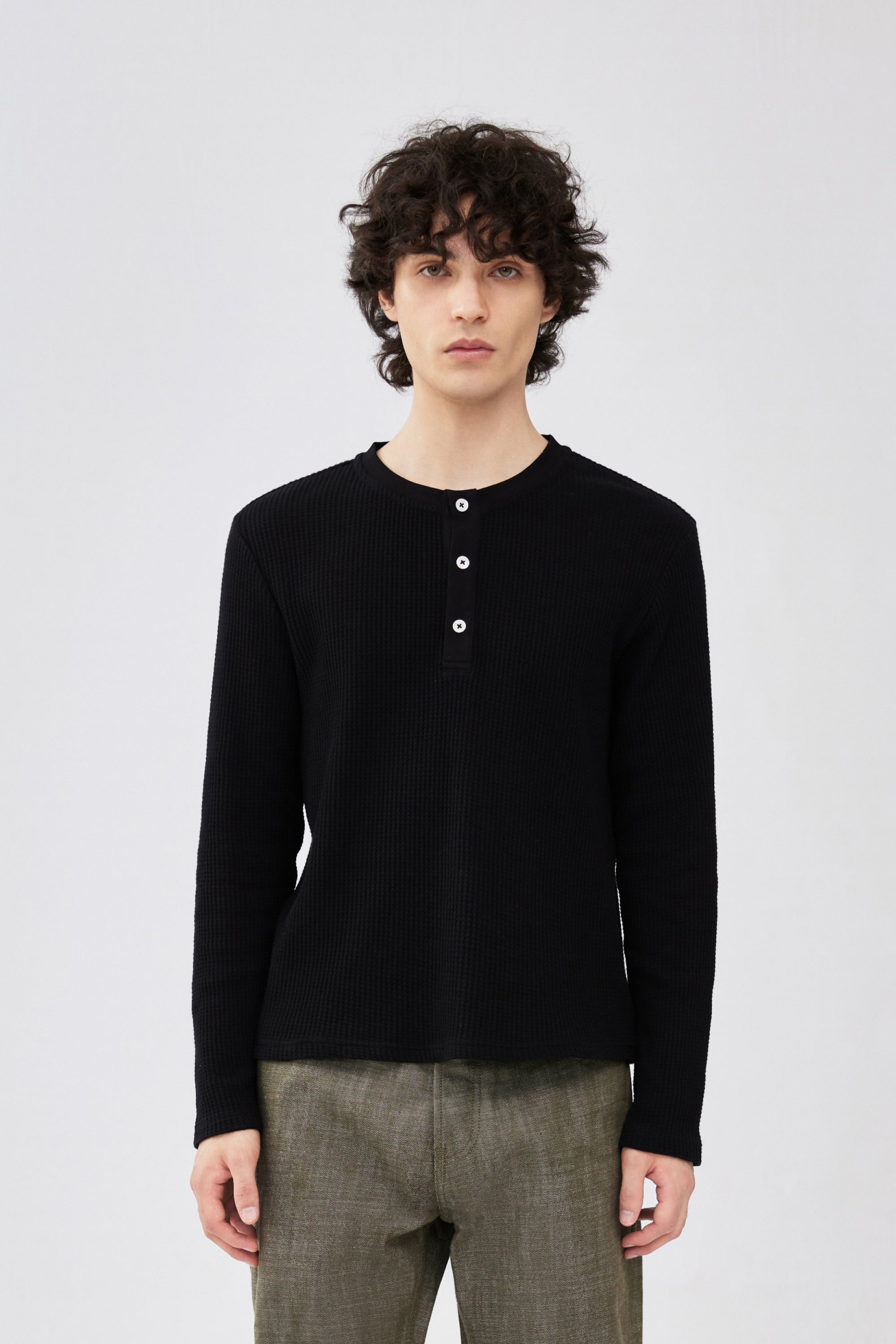 A male model wearing a black long-sleeve henley shirt with three white buttons and olive green trousers.