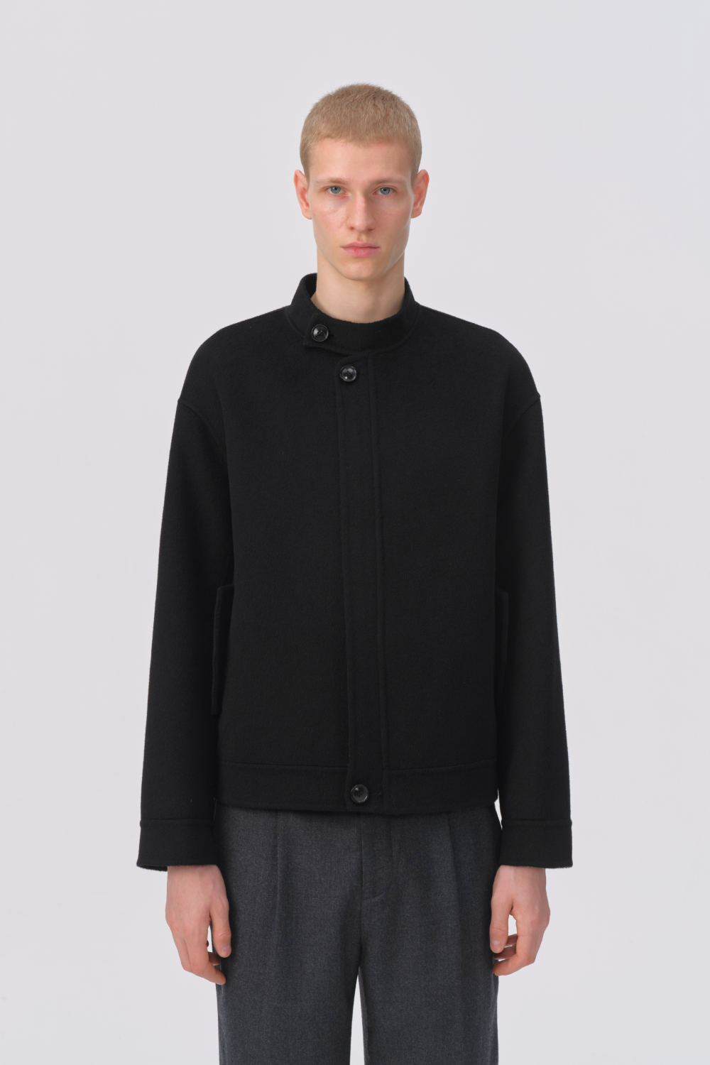 A male model wearing a black asymmetrical button-front jacket with a stand collar and dark gray trousers, standing against a light gray studio background.