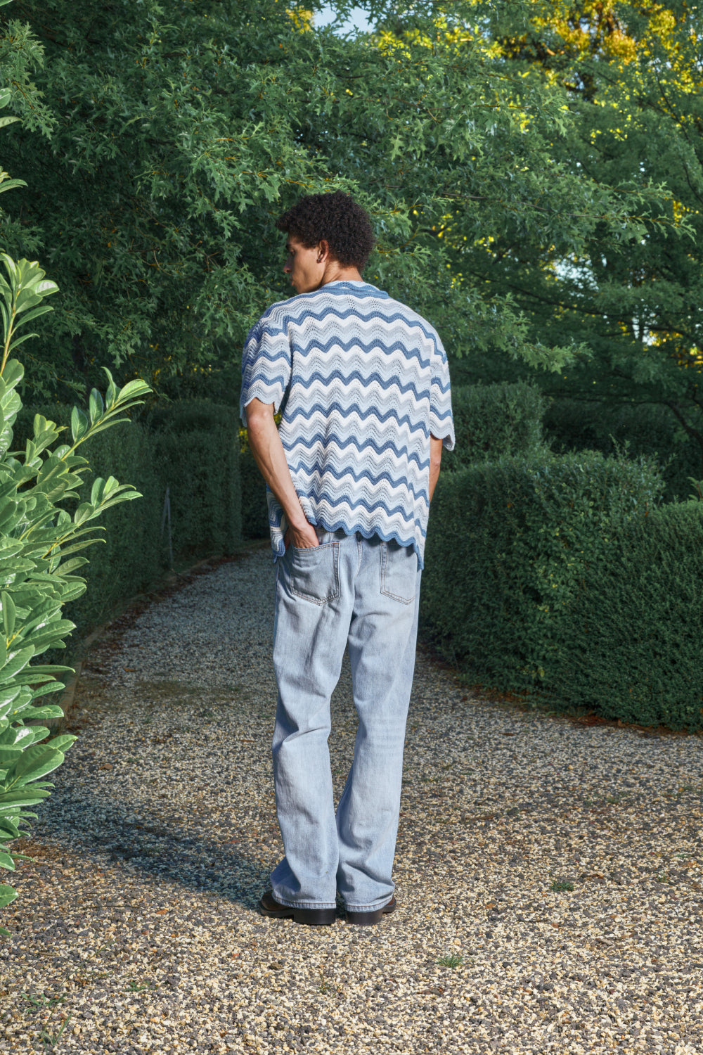 Back view for a model wearing a blue and white shale stitch knit shirt over a white top, standing outdoors.