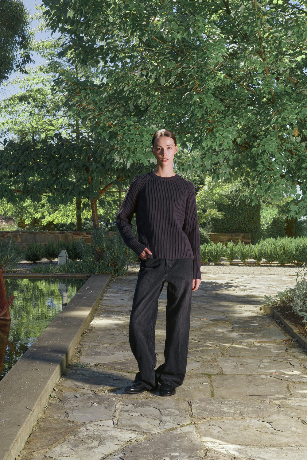 Model wearing a black ribbed knit sweater with black pants, standing outdoors with greenery in the background.