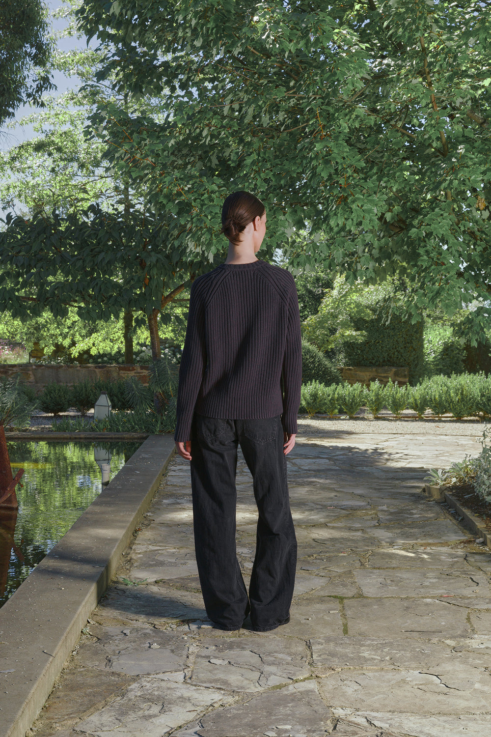 Full back view for a model wearing a black ribbed knit sweater with black pants, standing outdoors with greenery in the background.