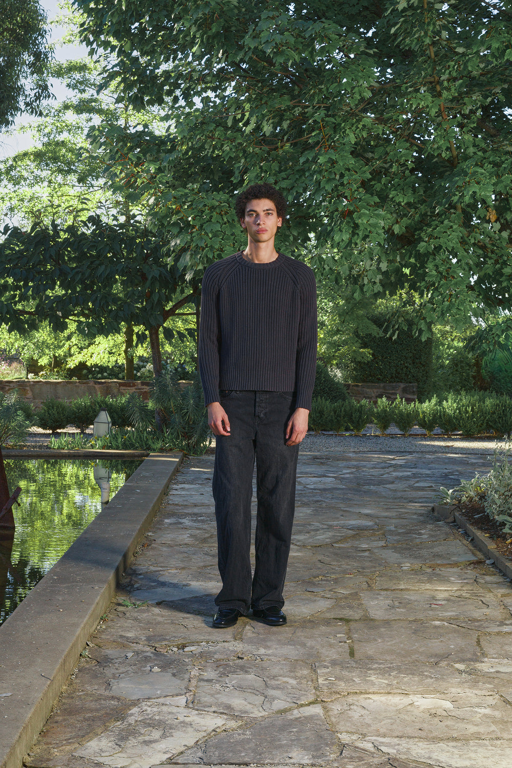 Model wearing a black ribbed knit sweater with black pants, standing on a stone pathway with greenery in the background