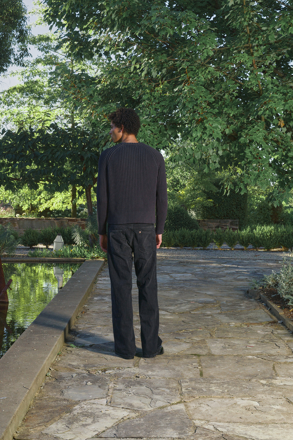 Back view for a model wearing a black ribbed knit sweater with black pants, standing outdoors.