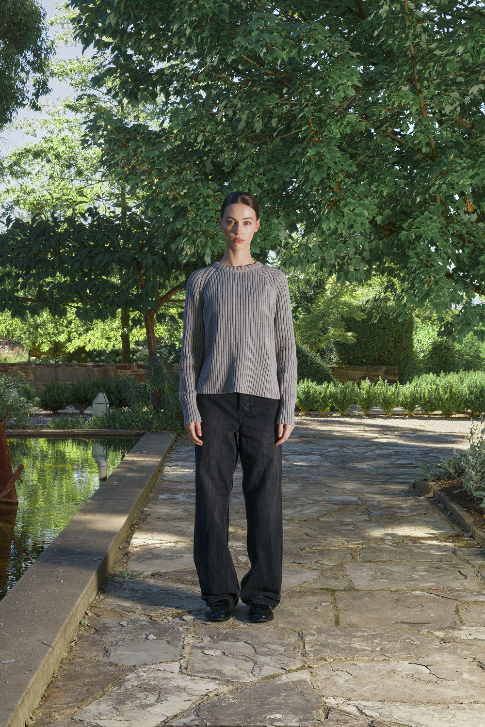 Model wearing a grey ribbed knit sweater with black pants, standing outdoors.
