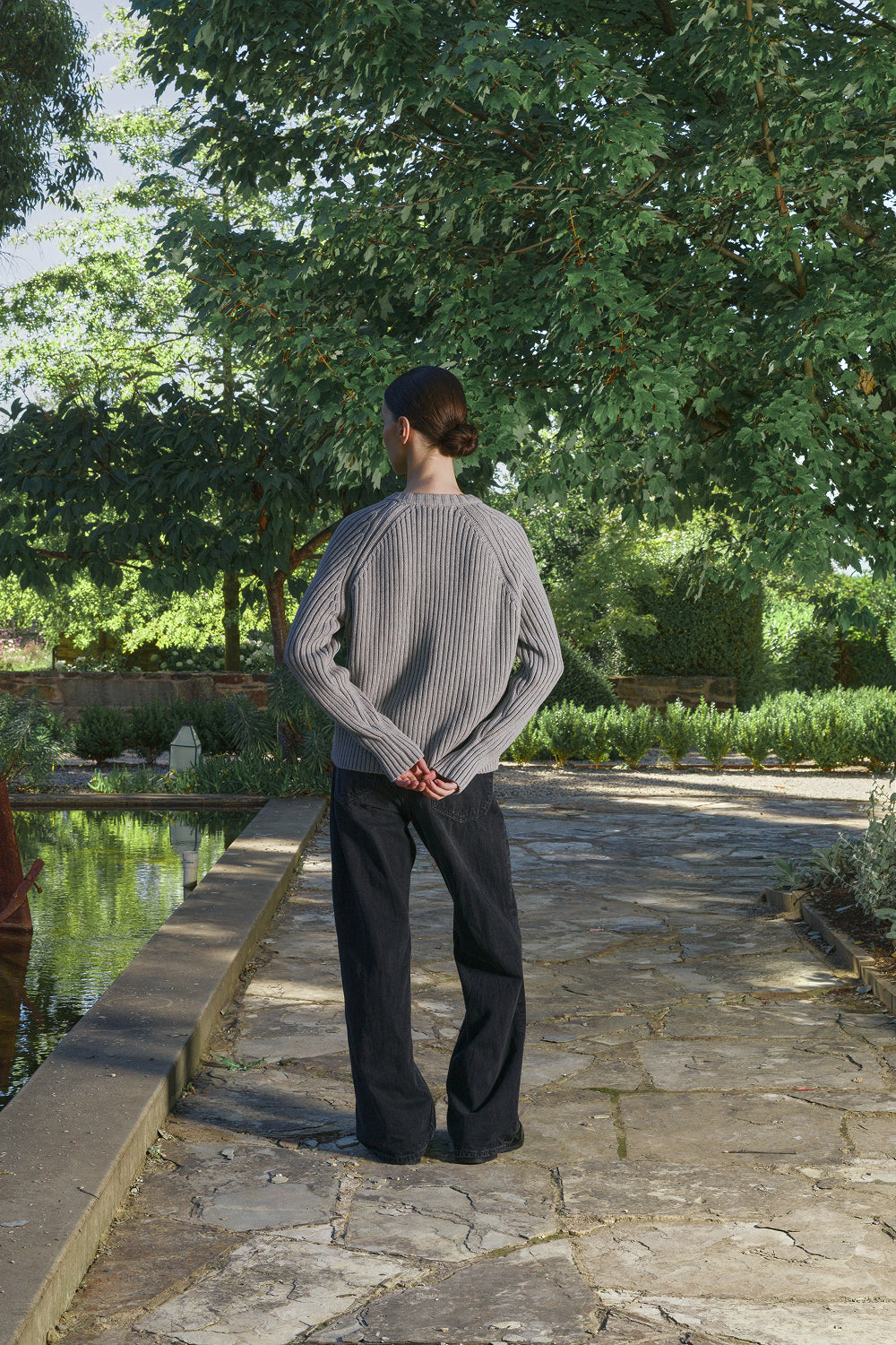 Back view for a model wearing a grey ribbed knit sweater with black pants, standing outdoors.