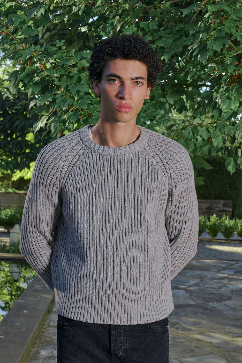 Close view for a model wearing a grey ribbed knit sweater with a black jeans, standing outdoors.
