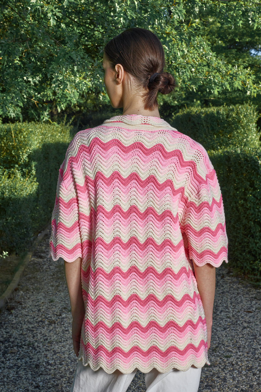 Back view of a woman wearing a pink and cream shale stitch crochet shirt, standing outdoors with greenery in the background.
