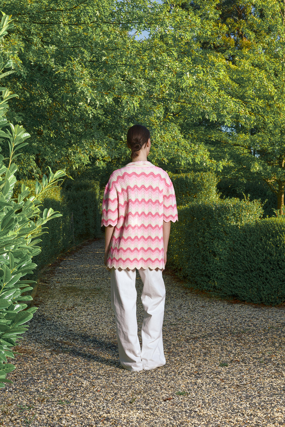 Back view of a woman wearing a pink and cream shale stitch crochet shirt with scalloped hem, standing on a garden path surrounded by greenery.