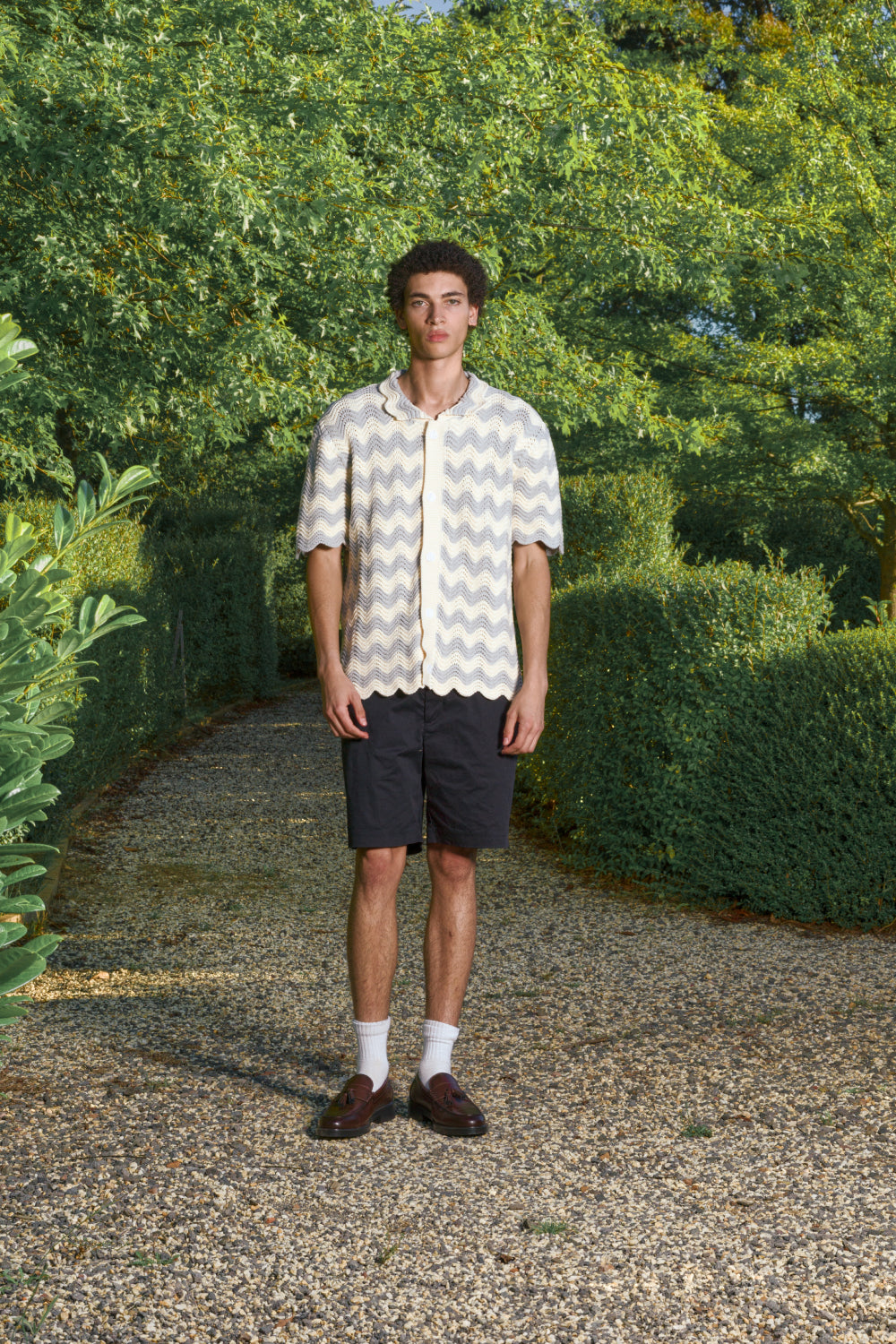 Man wearing a grey and cream shale stitch pattern shirt and black shorts standing in a garden.