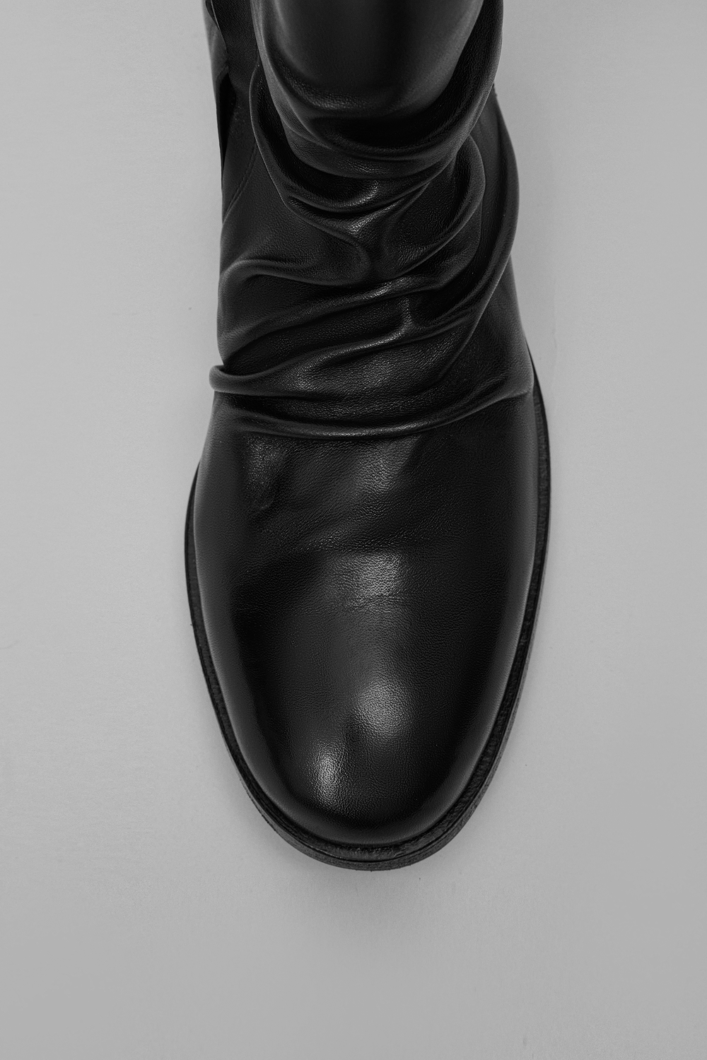 A close-up top-down view of a black leather boot with a draped, gathered shaft and rounded toe, set against a plain light gray background.