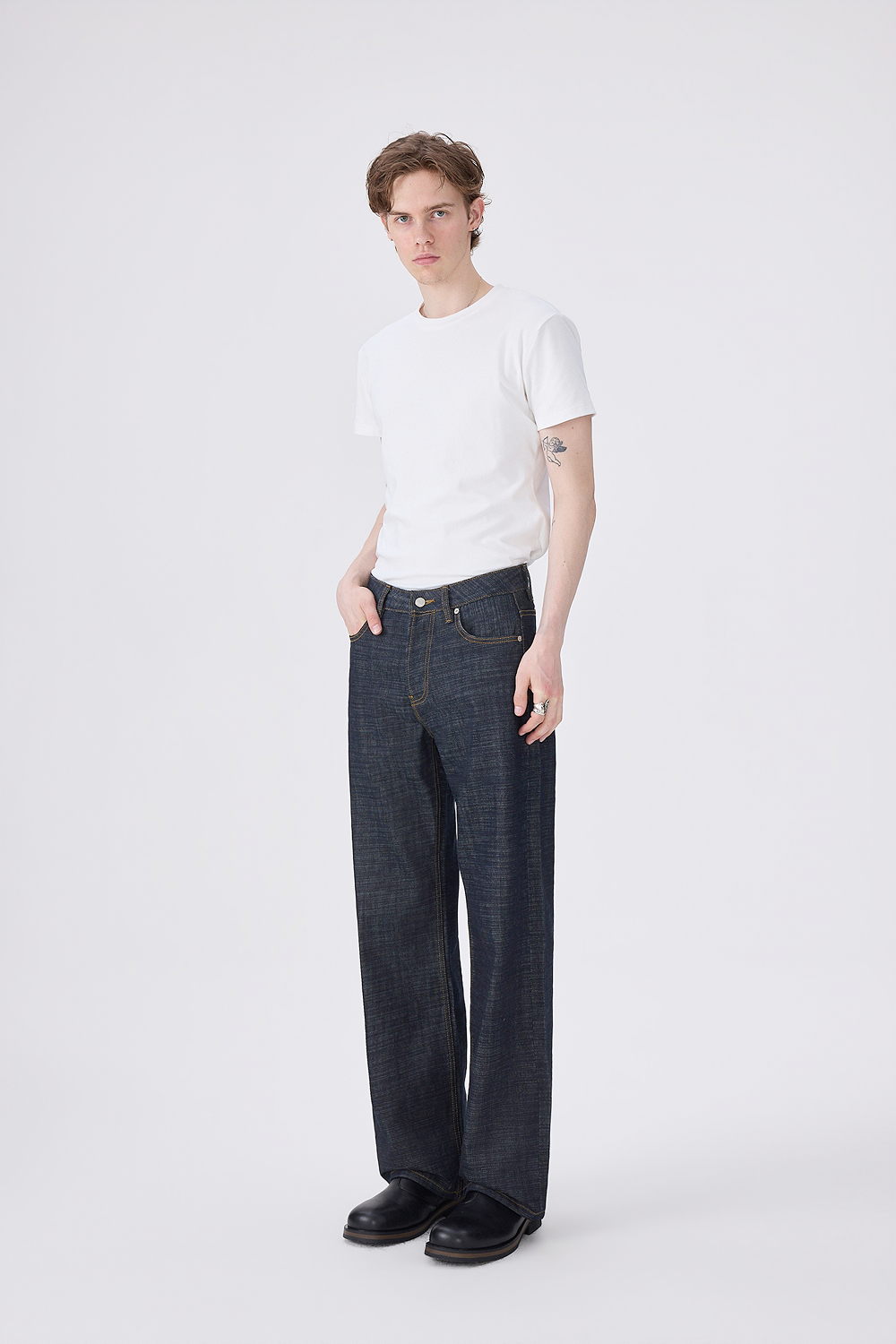 A model stands against a light gray studio background wearing a plain white short-sleeve T-shirt, dark indigo straight-leg jeans, and black shoes.