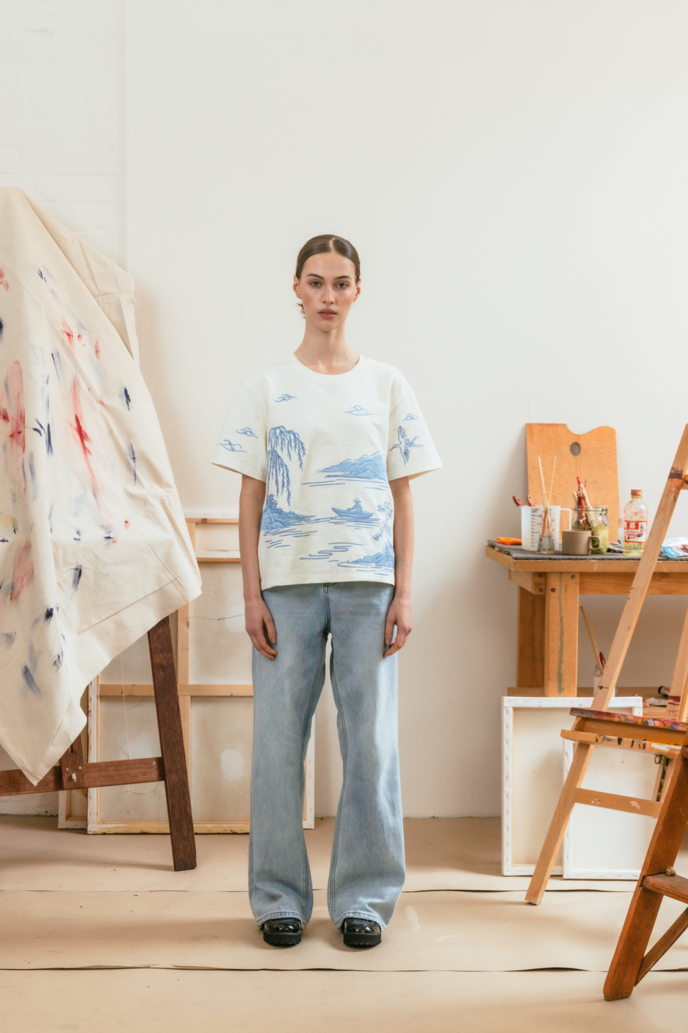 Person wearing a white t-shirt with blue embroidery and light blue jeans in an art studio.