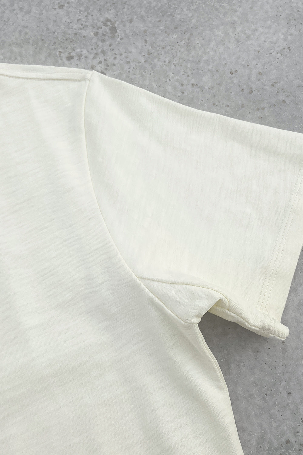 Close-up of the sleeve and side seam of a cream T-shirt laid flat on a gray textured surface, highlighting the soft fabric and stitching details.