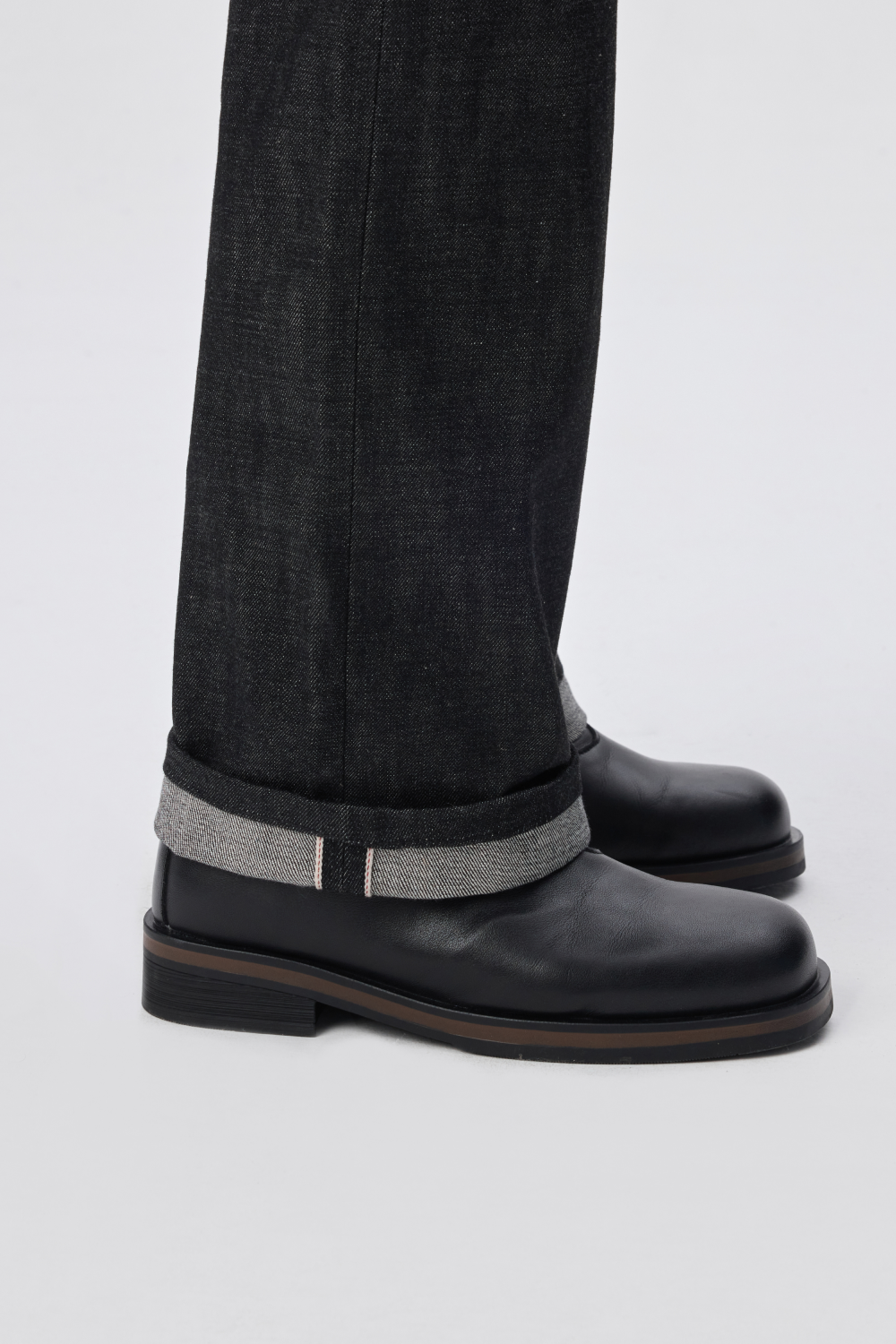 Close-up of black raw denim hem with a red selvedge edge, cuffed above black leather shoes with a stacked sole.