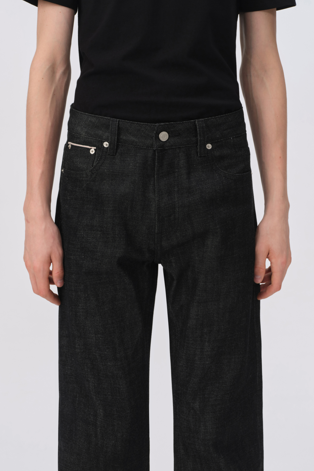Close-up of black raw denim waistband on a model, showing the button fly, coin pocket, and front pockets.
