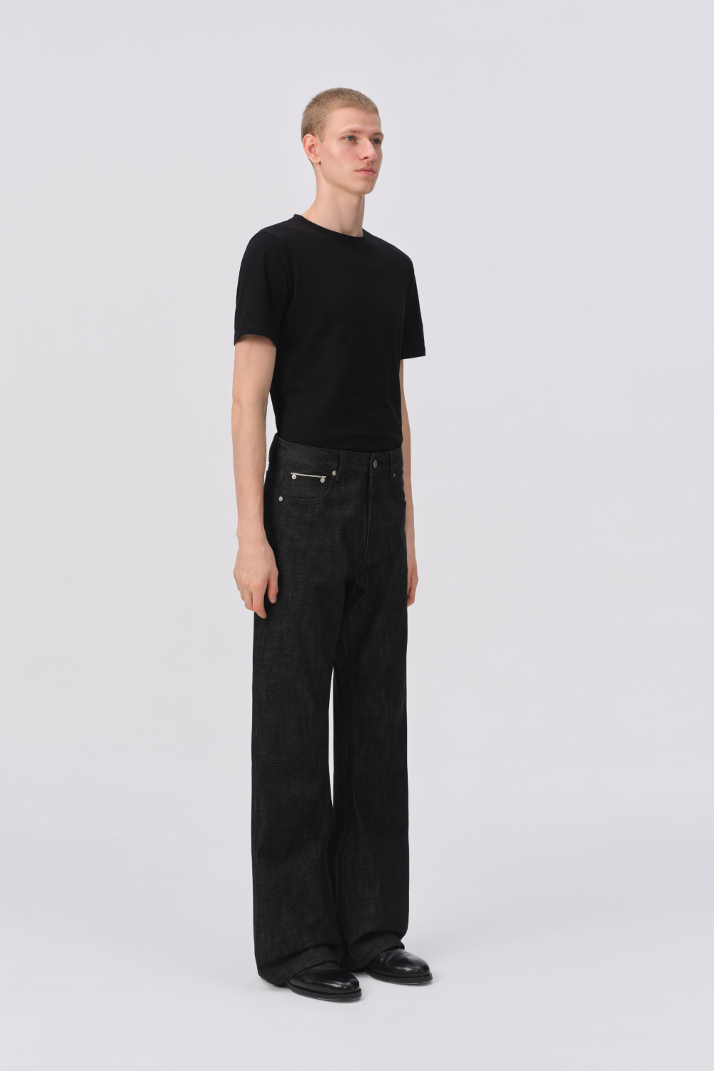 Full-length side view of a man in a black t-shirt and black raw denim straight-leg jeans styled with black shoes.
