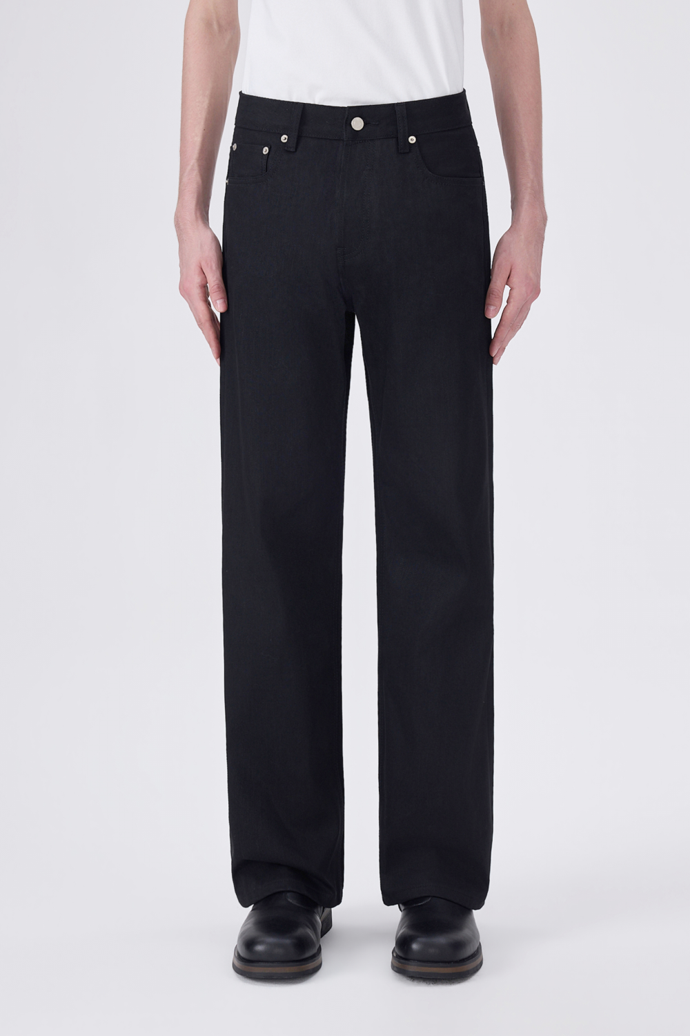 A model from the waist down wearing black straight-leg jeans, a white t-shirt, and black leather shoes, photographed against a light gray studio background.